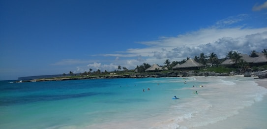 A picturesque beach scene featuring turquoise waters gently lapping against the white sandy shore. Thatched-roof huts and palm trees line the background under a blue sky with scattered clouds. People can be seen enjoying the water and relaxing along the shore.