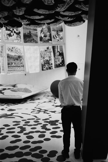 A person standing in an art gallery with black and white patterned flooring. The walls display various artworks or posters with diverse themes. The ceiling features abstract black shapes, and there is a large spherical object on the floor.