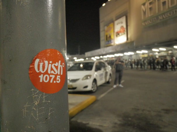 A sticker labeled 'Wish 107.5' is affixed to a gray pole in a nighttime setting. In the background, there is a parking lot with a white car and a crowd of people near the entrance of a building that has visible signage and lights.