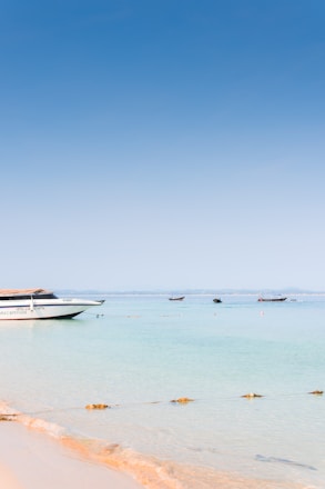 A serene beach scene with a small boat docked near crystal-clear water under a bright blue sky.