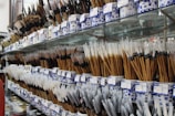 A variety of traditional calligraphy or art brushes are displayed on shelves, arranged in ornate blue and white ceramic holders. The brushes have bamboo handles and varied bristle colors and shapes, with price tags attached to each holder.