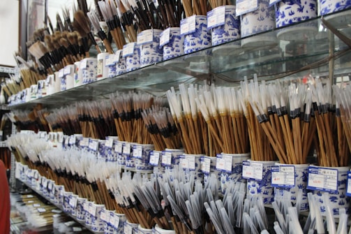 A variety of traditional calligraphy or art brushes are displayed on shelves, arranged in ornate blue and white ceramic holders. The brushes have bamboo handles and varied bristle colors and shapes, with price tags attached to each holder.