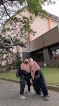 Two people wearing hijabs and casual clothing stand closely together on a paved path. They are smiling at the camera. In the background, there is a multi-story building with windows and a staircase, surrounded by lush green trees and grass.