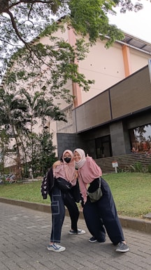 Two people wearing hijabs and casual clothing stand closely together on a paved path. They are smiling at the camera. In the background, there is a multi-story building with windows and a staircase, surrounded by lush green trees and grass.