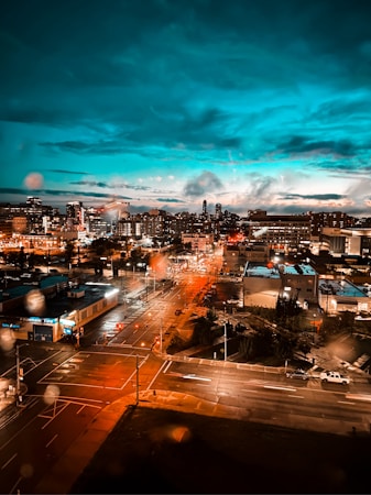 A vibrant cityscape at dusk with illuminated buildings, streetlights, and vehicles on roads. The sky features rich blue hues with scattered clouds, while the city lights create a warm, glowing effect. The image appears to have been taken from an elevated vantage point, capturing a wide view of urban activity and architecture. Raindrops on the camera lens add a subtle texture.