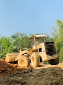 A large yellow bulldozer is positioned on a construction site, moving a pile of dirt. The background features lush green trees and a clear blue sky, indicating a rural or semi-rural setting. The ground appears to be uneven and consists of loose soil and rocks.
