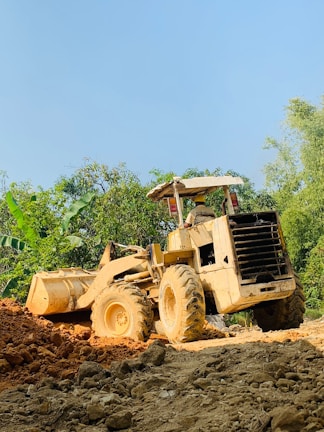 A large yellow bulldozer is positioned on a construction site, moving a pile of dirt. The background features lush green trees and a clear blue sky, indicating a rural or semi-rural setting. The ground appears to be uneven and consists of loose soil and rocks.