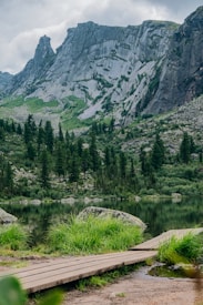 A serene mountain landscape featuring towering rocky cliffs with a backdrop of lush green vegetation. A calm lake reflects the surrounding scenery, while a wooden path leads to the water’s edge. Pine trees are scattered throughout the scene, enhancing the natural beauty.