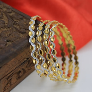 Several intricately designed gold and silver bangles are elegantly draped over an ornately carved wooden box. In the background, a soft red fabric provides a rich contrast.