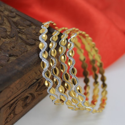 Several intricately designed gold and silver bangles are elegantly draped over an ornately carved wooden box. In the background, a soft red fabric provides a rich contrast.