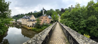 A stone pathway flanked by lush greenery extends into the distance. On one side, historic buildings with a mix of stone and stucco facades rest alongside a calm, reflective river. The setting feels serene and offers a picturesque view of nature harmoniously blending with architecture.