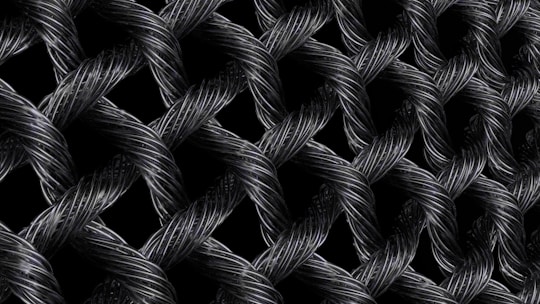 Interwoven strands of dark, metallic wires form a textured, lattice-like pattern. The intricate braiding creates a sense of depth and complexity, with highlights emphasizing the reflective surface of the materials.