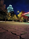 A nighttime scene featuring an illuminated, historic building with intricate architectural details amidst surrounding trees. A gas station with the sign 'TotalEnergies' glows in the foreground, alongside a brightly lit streetlamp. The ground is paved with textured, patterned stones.