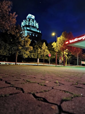 A nighttime scene featuring an illuminated, historic building with intricate architectural details amidst surrounding trees. A gas station with the sign 'TotalEnergies' glows in the foreground, alongside a brightly lit streetlamp. The ground is paved with textured, patterned stones.