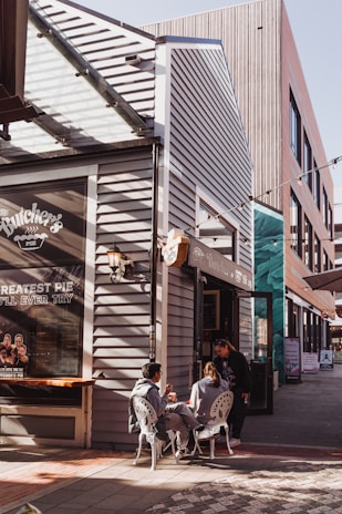 A small outdoor seating area with three people near a shop with a sign that reads 'The Butcher's Pie.' The design of the building features modern architectural elements with stylish fixtures and a display window promoting pies. Two individuals are seated at a table, while another is standing, creating a cozy and relaxed atmosphere.