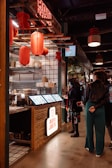 A modern food stall named Roti Joe's features a rustic wooden counter with digital menu screens. Red lanterns hang from the ceiling, providing warm lighting. Two people stand in front of the counter, engaging with the menu displays.