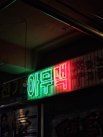 A neon sign with green and red Korean characters is mounted on a building exterior, casting a vibrant glow in an otherwise dimly lit urban setting. The sign contrasts with the dark surroundings, highlighting a gritty and atmospheric scene.