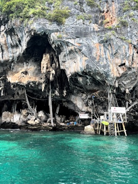 A rugged rock face with natural caves is seen above turquoise water. There are makeshift wooden platforms and scaffolding structures built against the cliffs. Some greenery grows on top of the rock formations.