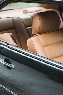 A close-up view of the interior of an elegant car, featuring luxurious tan leather seats. The focus is on the front seat with its soft, cushiony texture, highlighted by natural light. Part of the black exterior of the car's door is visible, emphasizing the contrast between the dark exterior and the light, inviting interior.