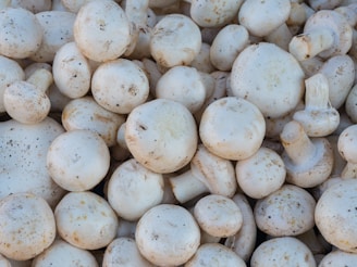 A collection of fresh white button mushrooms piled closely together, displaying a natural earthy texture with some speckles of soil visible on their surfaces.
