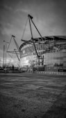 The image depicts a modern stadium with a sleek architectural design featuring large glass panels and distinctive steel cables supporting the structure. The foreground includes an empty paved area and several street lamps.