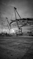 The image depicts a modern stadium with a sleek architectural design featuring large glass panels and distinctive steel cables supporting the structure. The foreground includes an empty paved area and several street lamps.