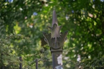 A wrought iron fence post topped with a decorative finial shaped like a blooming flower, surrounded by lush greenery in a garden setting. The background is softly blurred, highlighting the intricate details of the finial against the vibrant green foliage.