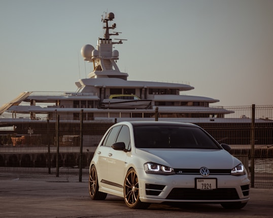 A white Volkswagen car is parked in front of a large yacht, with a metal fence separating them. The car is positioned slightly to the right, featuring black rims and prominent headlights. The yacht, visible in the background, is equipped with various antennae and radome structures, indicating its advanced technology. The sky appears clear, giving the scene a warm and serene evening ambiance.