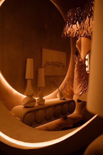 Warm ambient lighting illuminates a stylish interior with a modern console table featuring circular designs. Two tall vases sit atop the console, along with a small branch arrangement in a vase. An abstract art piece is mounted on the wall behind. A unique woven lampshade casts intriguing shadows.