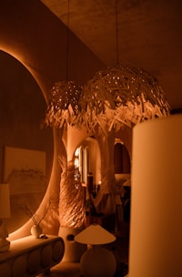 Warm ambient lighting fills a room decorated with stylish, organic-shaped furniture and accent pieces. Hanging woven light fixtures create a cozy atmosphere, with matching sculptures and vases enhancing the design. Archways and soft shadows add depth and intrigue to the setting.