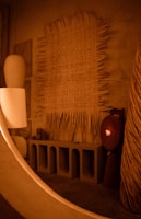 An interior scene featuring a variety of artistic and design elements. A large woven tapestry with fringes is mounted on a textured wall. Below it, a minimalist concrete console table with arched compartments holds several cylindrical and rectangular decorative objects. The ambient lighting is warm, contributing to a cozy atmosphere.