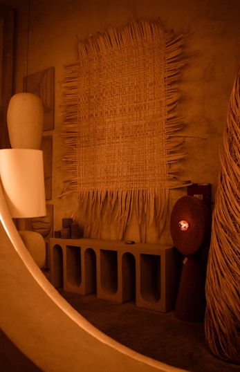 An interior scene featuring a variety of artistic and design elements. A large woven tapestry with fringes is mounted on a textured wall. Below it, a minimalist concrete console table with arched compartments holds several cylindrical and rectangular decorative objects. The ambient lighting is warm, contributing to a cozy atmosphere.