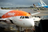 A commercial airplane from easyJet is stationed at an airport gate with a name 'Ayshe Ballard' visible on the side. The aircraft is parked on the tarmac, with its nose facing the camera. A set of mobile stairs with 'Menzies' branding is positioned at the passenger door, alongside airport personnel. In the background, other airplanes from KLM and TUI are visible, parked on a wet runway.