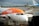 A commercial airplane from easyJet is stationed at an airport gate with a name 'Ayshe Ballard' visible on the side. The aircraft is parked on the tarmac, with its nose facing the camera. A set of mobile stairs with 'Menzies' branding is positioned at the passenger door, alongside airport personnel. In the background, other airplanes from KLM and TUI are visible, parked on a wet runway.