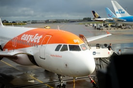 A commercial airplane from easyJet is stationed at an airport gate with a name 'Ayshe Ballard' visible on the side. The aircraft is parked on the tarmac, with its nose facing the camera. A set of mobile stairs with 'Menzies' branding is positioned at the passenger door, alongside airport personnel. In the background, other airplanes from KLM and TUI are visible, parked on a wet runway.