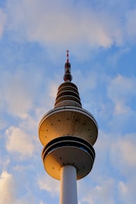 A tall, cylindrical telecommunications tower with multiple observation decks and a red-and-white antenna at the top stands against a backdrop of a blue sky with scattered clouds.