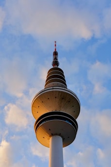 A tall, cylindrical telecommunications tower with multiple observation decks and a red-and-white antenna at the top stands against a backdrop of a blue sky with scattered clouds.