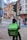 A delivery person on a bicycle waits at a traffic light with a large green Uber Eats backpack. The urban setting features historic and modern buildings, a 20 mph speed limit sign, and another cyclist can be seen in the background, creating a balance of traditional architecture with contemporary lifestyle.
