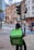 A delivery person on a bicycle waits at a traffic light with a large green Uber Eats backpack. The urban setting features historic and modern buildings, a 20 mph speed limit sign, and another cyclist can be seen in the background, creating a balance of traditional architecture with contemporary lifestyle.