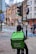 A delivery person on a bicycle waits at a traffic light with a large green Uber Eats backpack. The urban setting features historic and modern buildings, a 20 mph speed limit sign, and another cyclist can be seen in the background, creating a balance of traditional architecture with contemporary lifestyle.