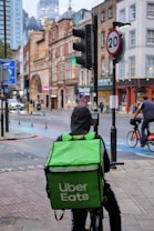 A delivery person on a bicycle waits at a traffic light with a large green Uber Eats backpack. The urban setting features historic and modern buildings, a 20 mph speed limit sign, and another cyclist can be seen in the background, creating a balance of traditional architecture with contemporary lifestyle.