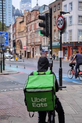 A delivery person on a bicycle waits at a traffic light with a large green Uber Eats backpack. The urban setting features historic and modern buildings, a 20 mph speed limit sign, and another cyclist can be seen in the background, creating a balance of traditional architecture with contemporary lifestyle.