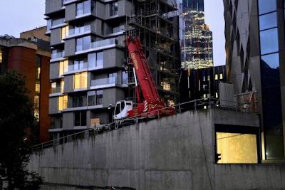 A modern cityscape features a construction site with a prominent red crane situated in front of a building with large glass windows. Adjacent buildings have diverse architectural styles, including a high-rise with reflective glass. Evening light casts a subdued glow.