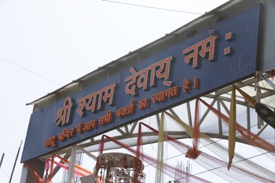 An entrance gate decorated with colorful fabric and adorned with Hindi script. The structure includes text in red and white on a blue background, with decorative elements like ribbons and a traditional hanging ornament underneath.