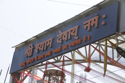 An entrance gate decorated with colorful fabric and adorned with Hindi script. The structure includes text in red and white on a blue background, with decorative elements like ribbons and a traditional hanging ornament underneath.