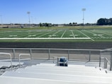 A sports field with lines marking football or soccer measurements is surrounded by a running track. Several tall floodlights are positioned around the area and empty metal bleachers can be seen in the foreground. In the distance, there are trees and buildings under a clear blue sky.