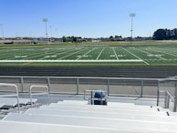 A sports field with lines marking football or soccer measurements is surrounded by a running track. Several tall floodlights are positioned around the area and empty metal bleachers can be seen in the foreground. In the distance, there are trees and buildings under a clear blue sky.