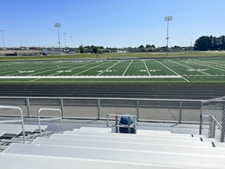 A sports field with lines marking football or soccer measurements is surrounded by a running track. Several tall floodlights are positioned around the area and empty metal bleachers can be seen in the foreground. In the distance, there are trees and buildings under a clear blue sky.