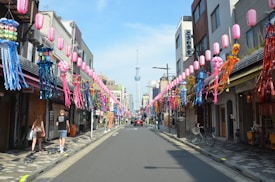 A festive street lined with colorful decorations, including vibrant streamers and lanterns. People walk along the sidewalks, and a red car is parked on the right side of the road. Traditional buildings and shops are visible on both sides, with a tower viewable in the background under a clear sky.