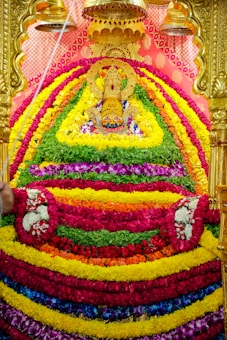 An ornate Hindu idol adorned with an intricate arrangement of colorful flower garlands. The idol is set against a decorative gold backdrop with bells hanging above. The vibrant garlands are laid in layers of red, yellow, green, orange, and pink, creating a striking visual display.